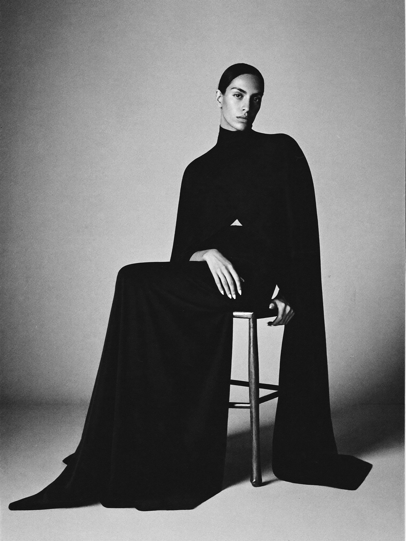 Seated study in sculptural black drape, fabric pooling on the floor, monochrome.