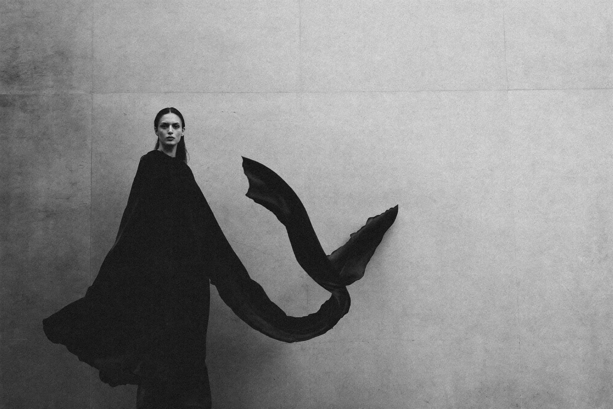 A model in a fluid black draped garment against a pale studio wall, monochrome editorial.