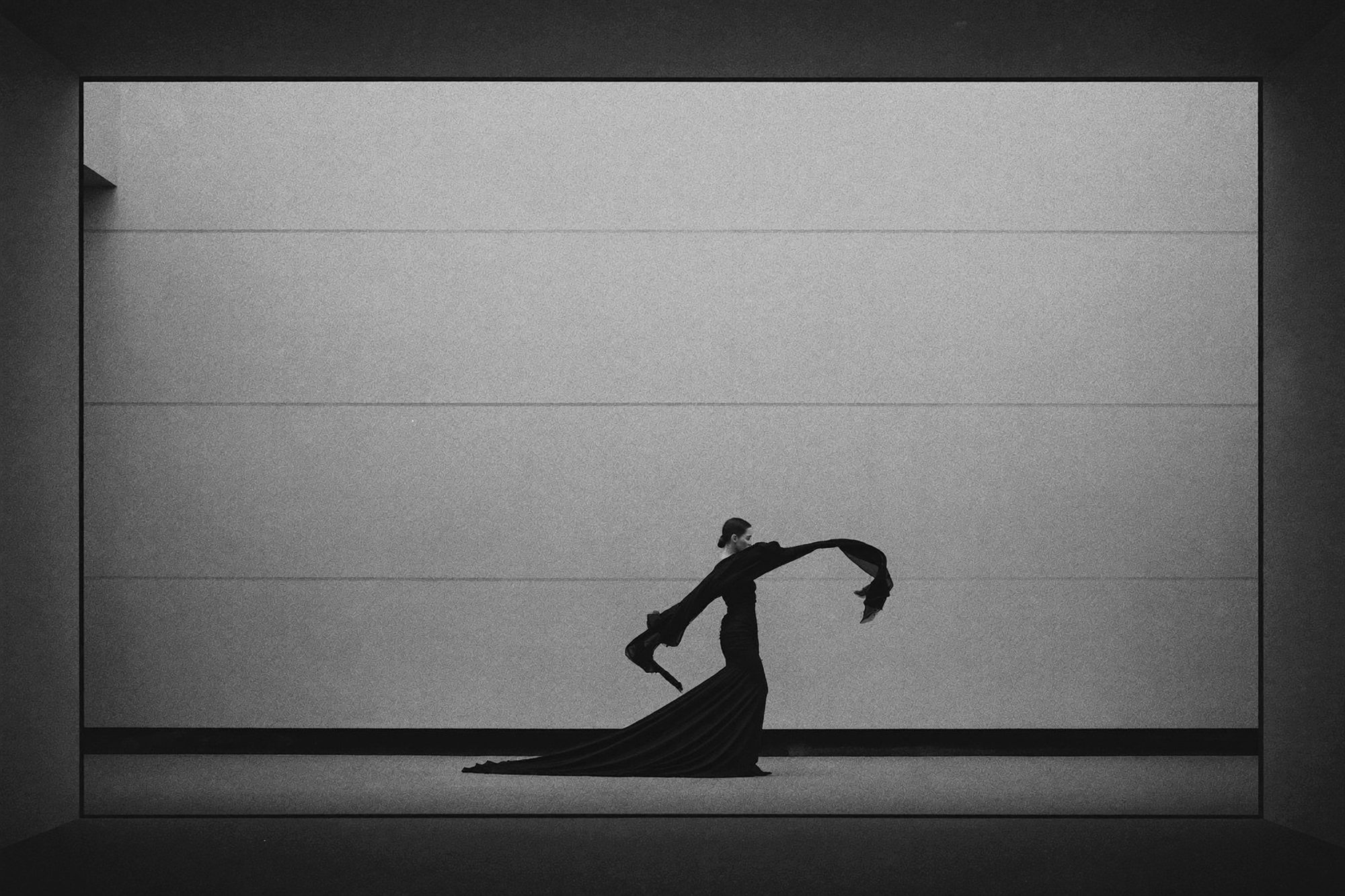 Wide monochrome editorial: a model in flowing black against an expanse of pale concrete.